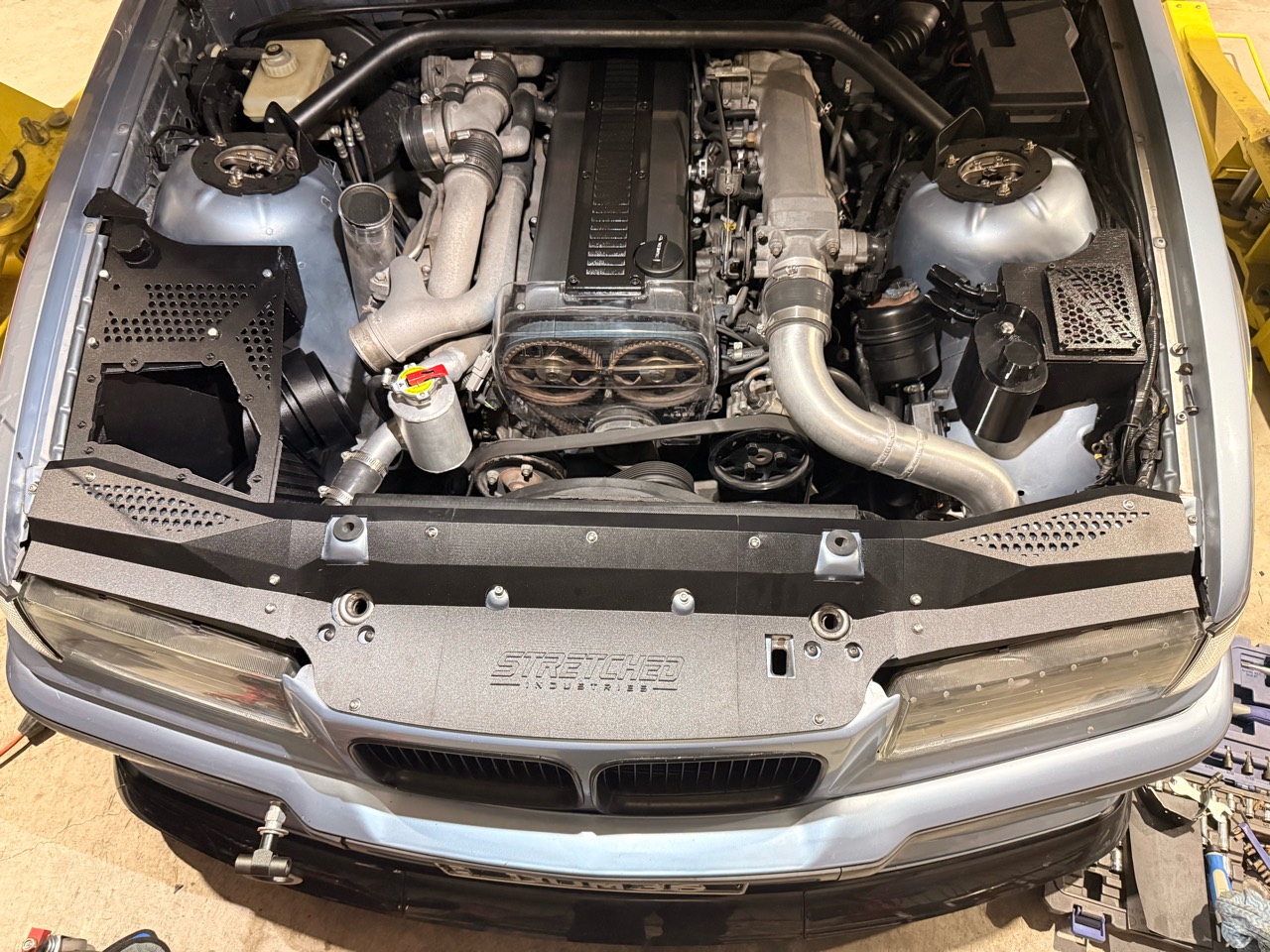 Front view into the E36 engine bay showing the Stretched Industries radiator support cover and surrounding development parts